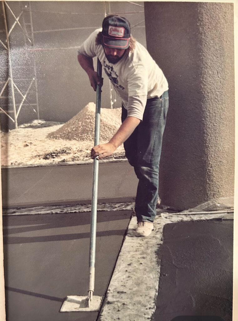 A Man in a grey hat and white shirt smooths out cement at a job site.