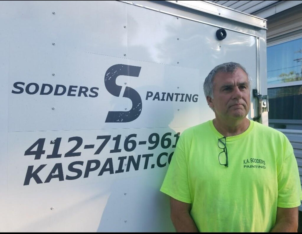 A man in a bright neon yellow shirt poses in front of the K.A. Sodders Logo.