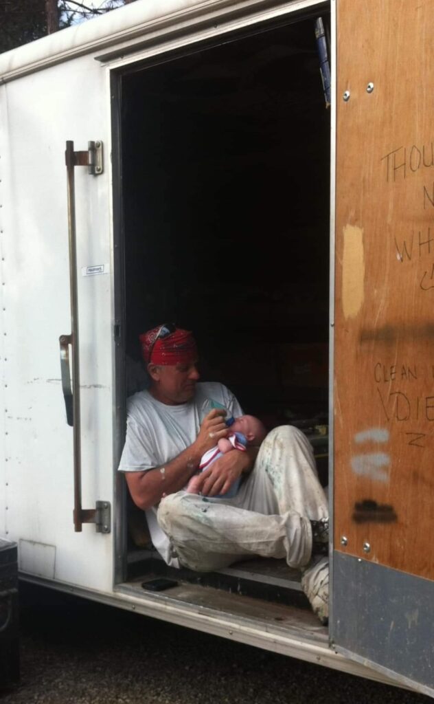 A man in a white shirt and pants sits in a storage trailer while holding and bottle-feeding a newborn baby.